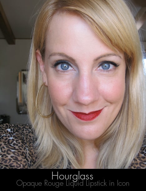 Een blonde vrouw die glimlachend de zandloper ondoorzichtige rouge vloeibare lippenstift draagt in icoon met een rijke blauwrode tint lippenstift