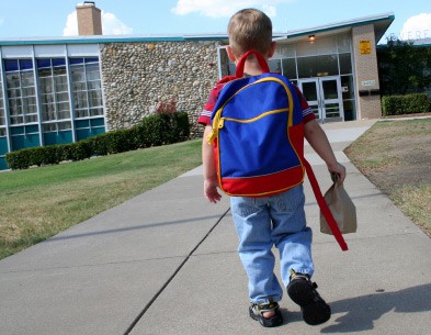 een kind dat loopt en een roodblauwe en gele rugzak draagt en een papieren tas draagt